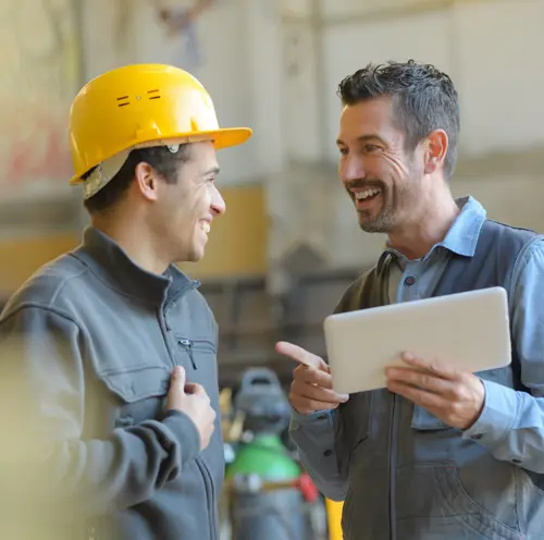 A couple of men standing next to each other in hard hats and overalls looking at a tablet.